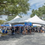 A large white tent with a blue banner reading “Falcons Forever Homecoming, Massage Therapy Program” shelters a crowd of people in gray and blue event shirts. Smaller blue canopies, a dog on a leash, and shady trees surround the parking lot under a partly cloudy sky.
