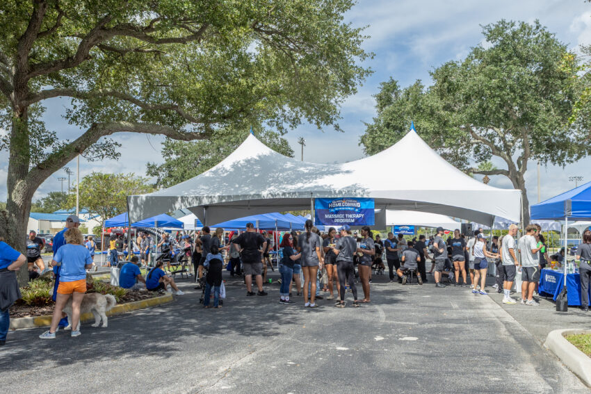 A large white tent with a blue banner reading “Falcons Forever Homecoming, Massage Therapy Program” shelters a crowd of people in gray and blue event shirts. Smaller blue canopies, a dog on a leash, and shady trees surround the parking lot under a partly cloudy sky.