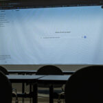 A dimly lit classroom with empty desks and chairs facing a projector screen displaying ChatGPT with the question "How Should AI Be Used In School?" typed in the prompt bar.