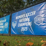 Two blue banners on a chain-link fence display Daytona State Women's Soccer as 2024 and 2025 NJCAA National Champions, with grass and fallen leaves in the foreground.