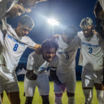 Boys' soccer players wearing white uniforms gather in a tight huddle with their heads bowed beneath a bright stadium light at night.