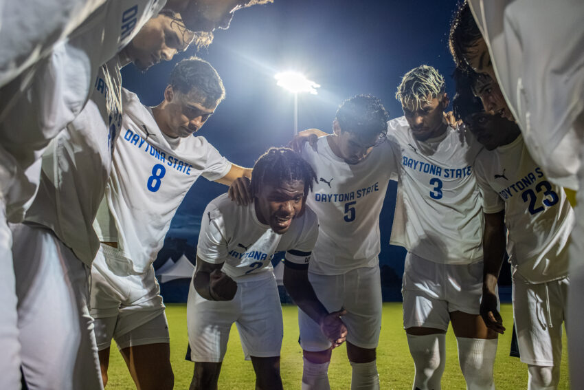 Boys' soccer players wearing white uniforms gather in a tight huddle with their heads bowed beneath a bright stadium light at night.