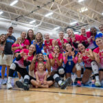 A large group of girl's volleyball players wearing pink jerseys pose together on an indoor court, smiling and raising their arms while coaches and teammates gather around them. All making Ws with their hands.
